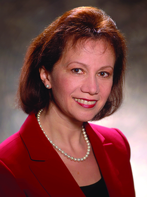 Headshot of Bella Goren, a 1983 graduate of the Chemical Engineering Department at UT Austin, wearing a red suit, black top and pearl necklace sitting in front of a gray background.